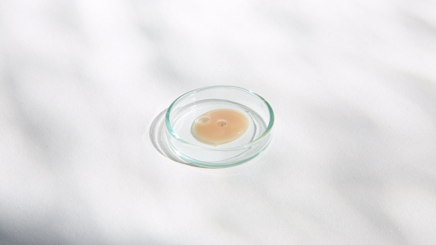 A small round glass Petri dish contains a circular drop of a beige-colored substance, 123 LActive face serum with CBD, placed on a white surface. Soft shadows create a subtle pattern in the background.