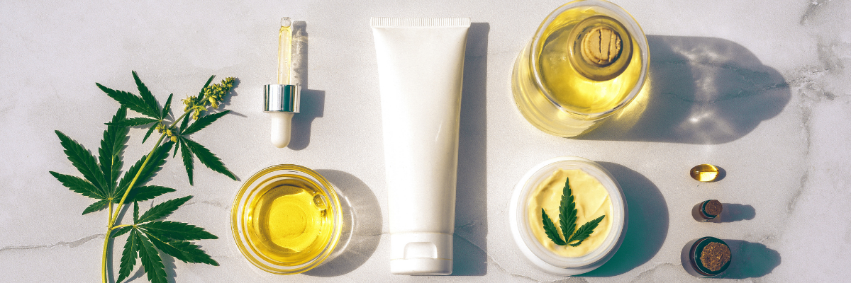 Divers produits de soins de la peau et de bien-être, notamment des pots, un tube, un compte-gouttes et des capsules, sont disposés sur une surface en marbre à côté d'une feuille de chanvre et d'huiles de cannabidiol (CBD) dans des récipients en verre.