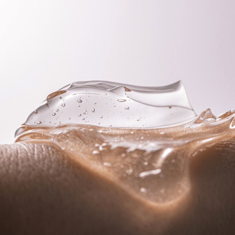 Gros plan d'un gel transparent avec de minuscules bulles d'air étalées sur une peau lisse et claire sur un fond doux et neutre.
