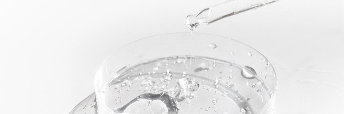 Un compte-gouttes déverse un liquide clair, riche en acide hyaluronique, dans une boîte de Petri remplie d'une substance transparente. De petites bulles dansent au sein du liquide dans la boîte, tandis que le fond blanc souligne la clarté et la simplicité de la scène.