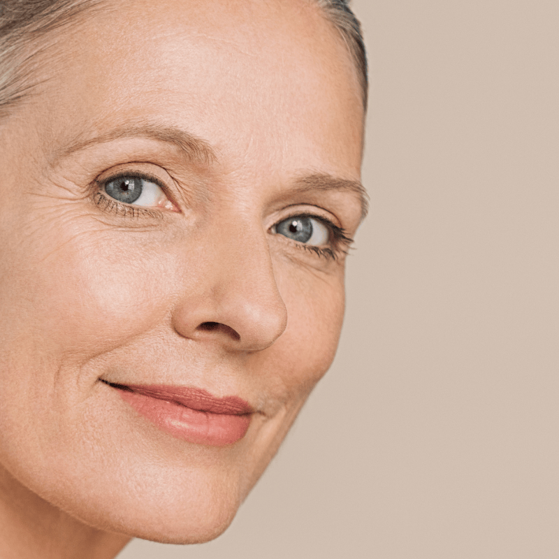 Visage d'une femme mature souriant et regardant l'objectif sur fond clair. Elle présente une peau vieillissante.