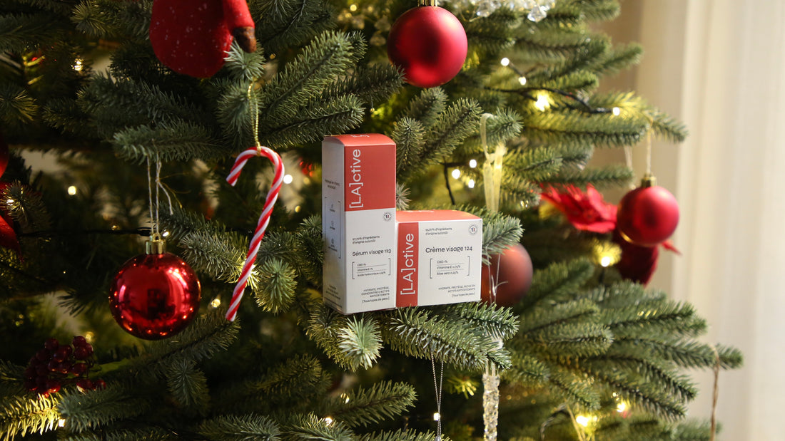 Un sapin de Noël décoré de boules rouges et blanches. Deux boîtes de produits de soin LActive contenant le sérum hydratant 123 et de la crème hydratante 124 sont nichées entre les branches.