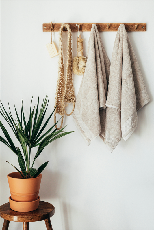 A wooden wall rack holds two beige towels, a small straw brush, and a mesh bath sponge. Below, a wooden stool supports a potted tropical plant with long, narrow green leaves. The wall behind is white, creating a clean and minimalist bathroom decor.