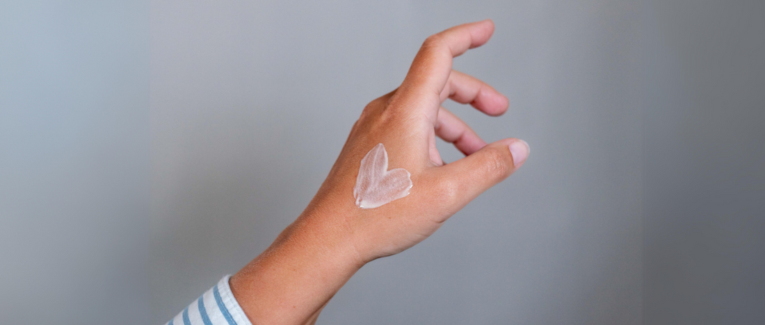 A hand with a heart-shaped dab of lotion on the back of it, against a plain background. The hand is slightly raised and positioned diagonally, wearing a striped long-sleeve shirt.