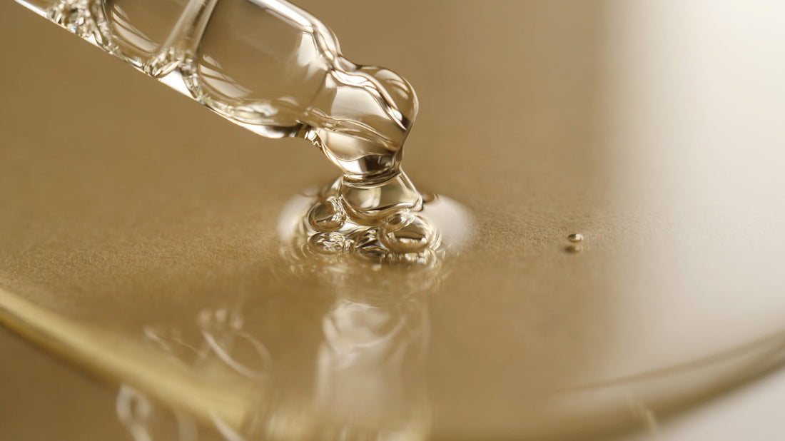A close-up of a clear liquid being dispensed from a glass dropper onto a golden surface, creating ripples. The liquid appears viscous and shiny, reflecting the light.