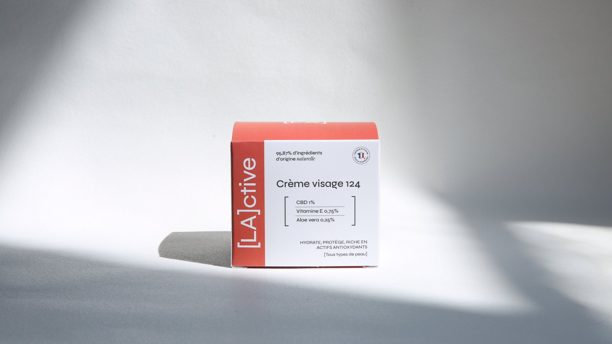 A can of LActive Crème Visage 124 is shown against a neutral background. The packaging states that it contains CBD, vitamin E, aloe vera and 95.87% ingredients of natural origin. The box is predominantly white, with text and branding elements in red and black. The box also states that the cream moisturizes, protects and is rich in antioxidant actives. Light and shadow play on the surface.