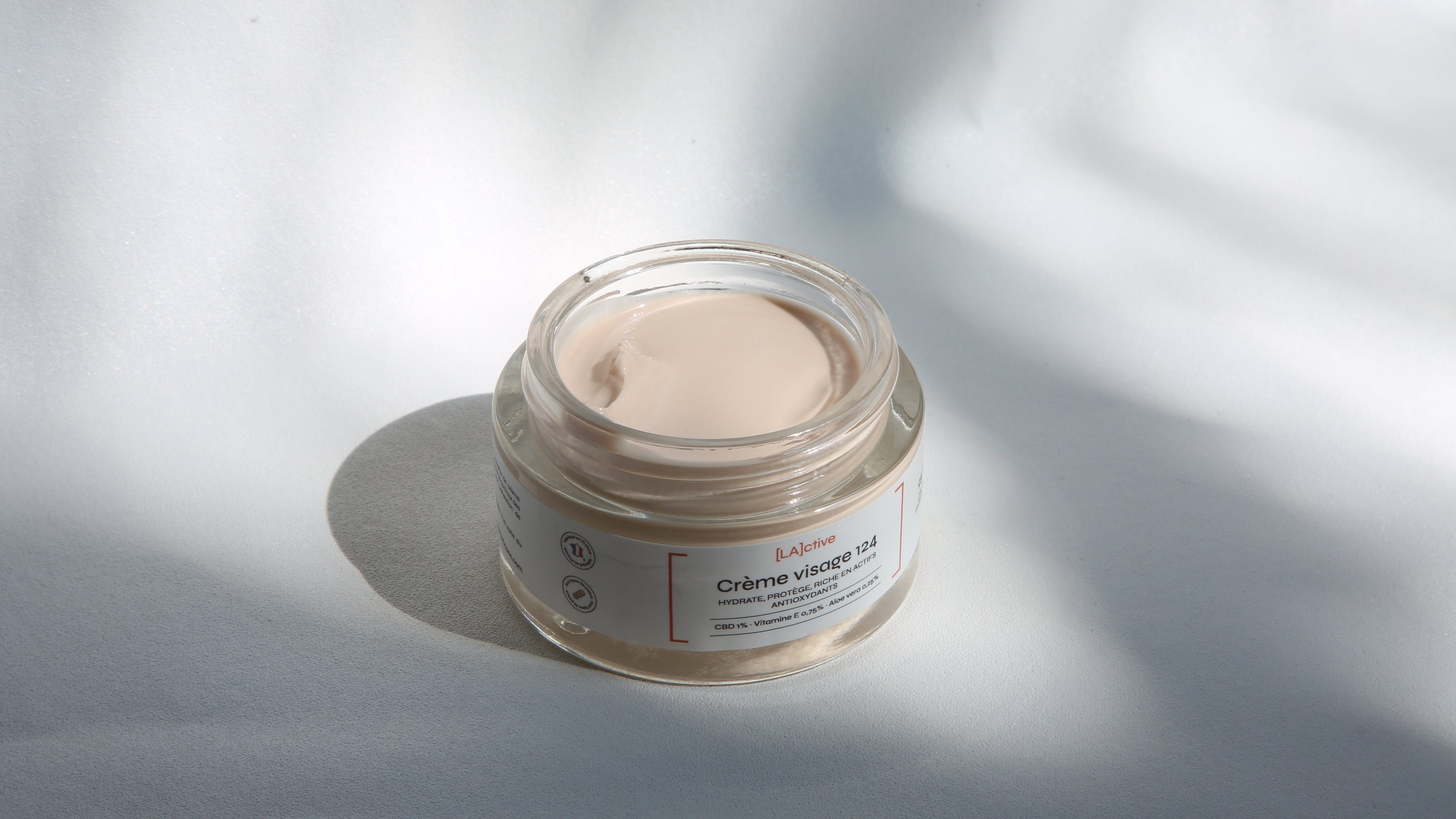 A jar of La Crème skincare cream, infused with CBD (cannabidiol), featuring a white label and light brown cream inside, rests on a smooth, white surface with soft shadows.