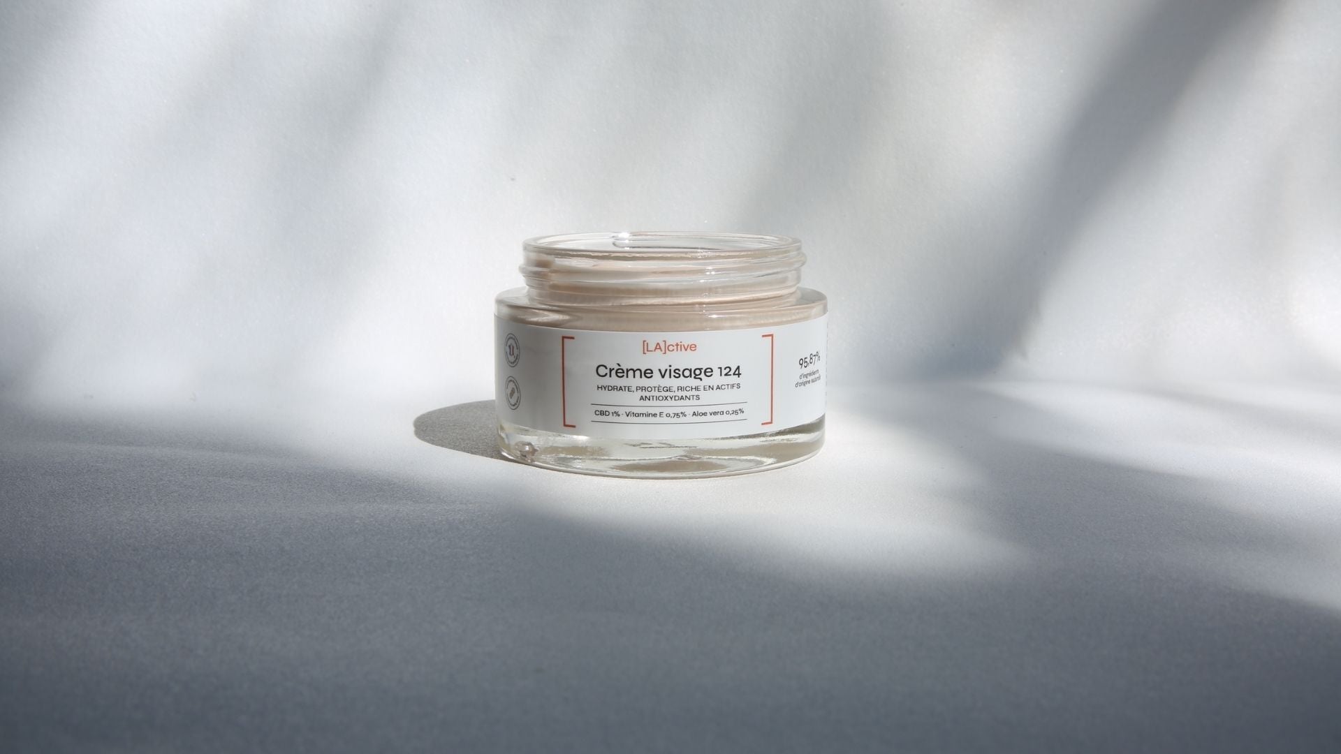 A jar of Crème Visage 124 is placed on a surface, with light casting shadows. French text highlights the cream's hydrating, protective, and illuminating properties.