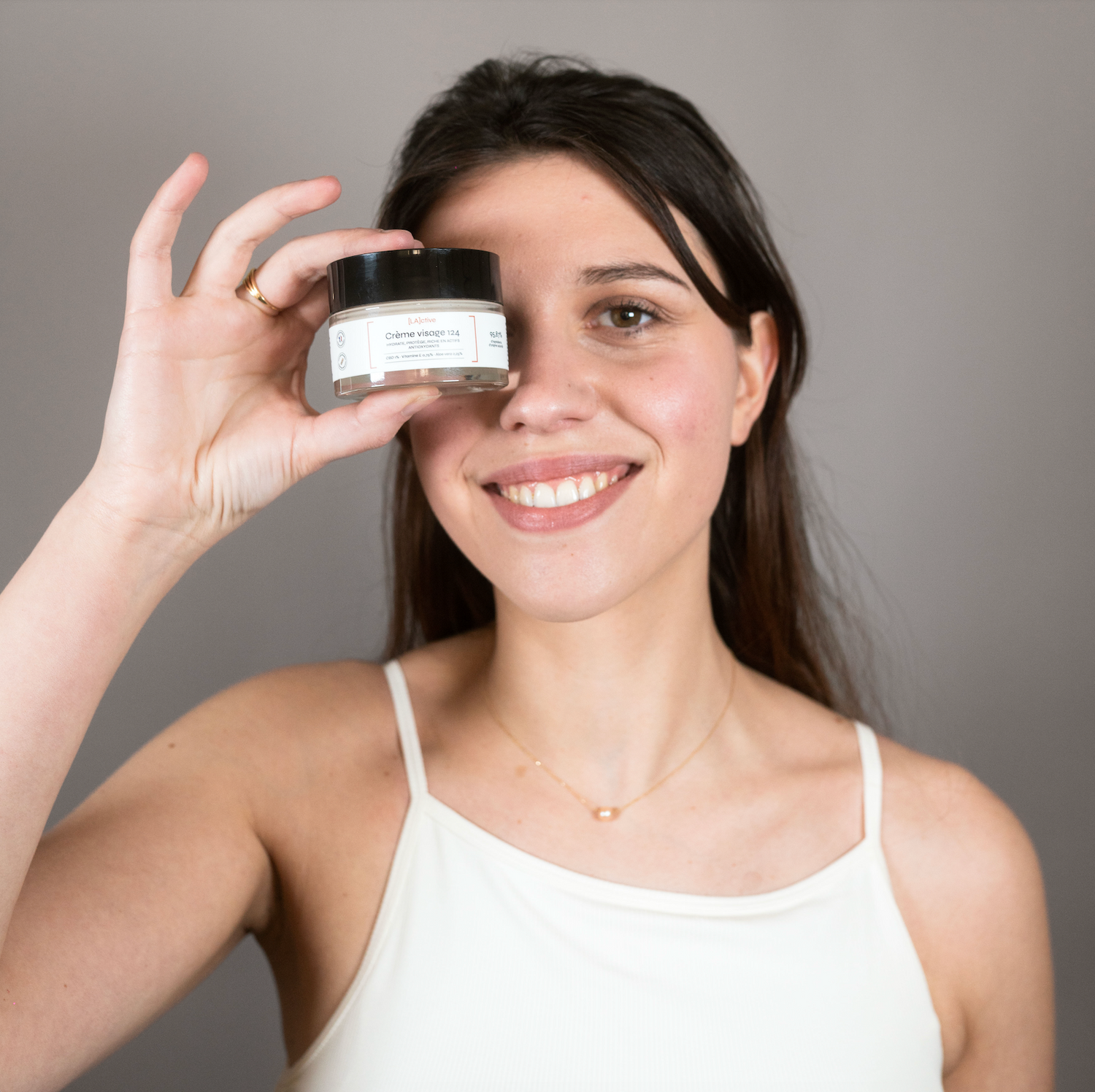 Une femme sourit tout en tenant un pot de crème hydratante visage lactive près de son visage, mettant en valeur l'essence de la cosmétique. Elle porte un débardeur blanc avec un collier simple sur un fond gris uni.