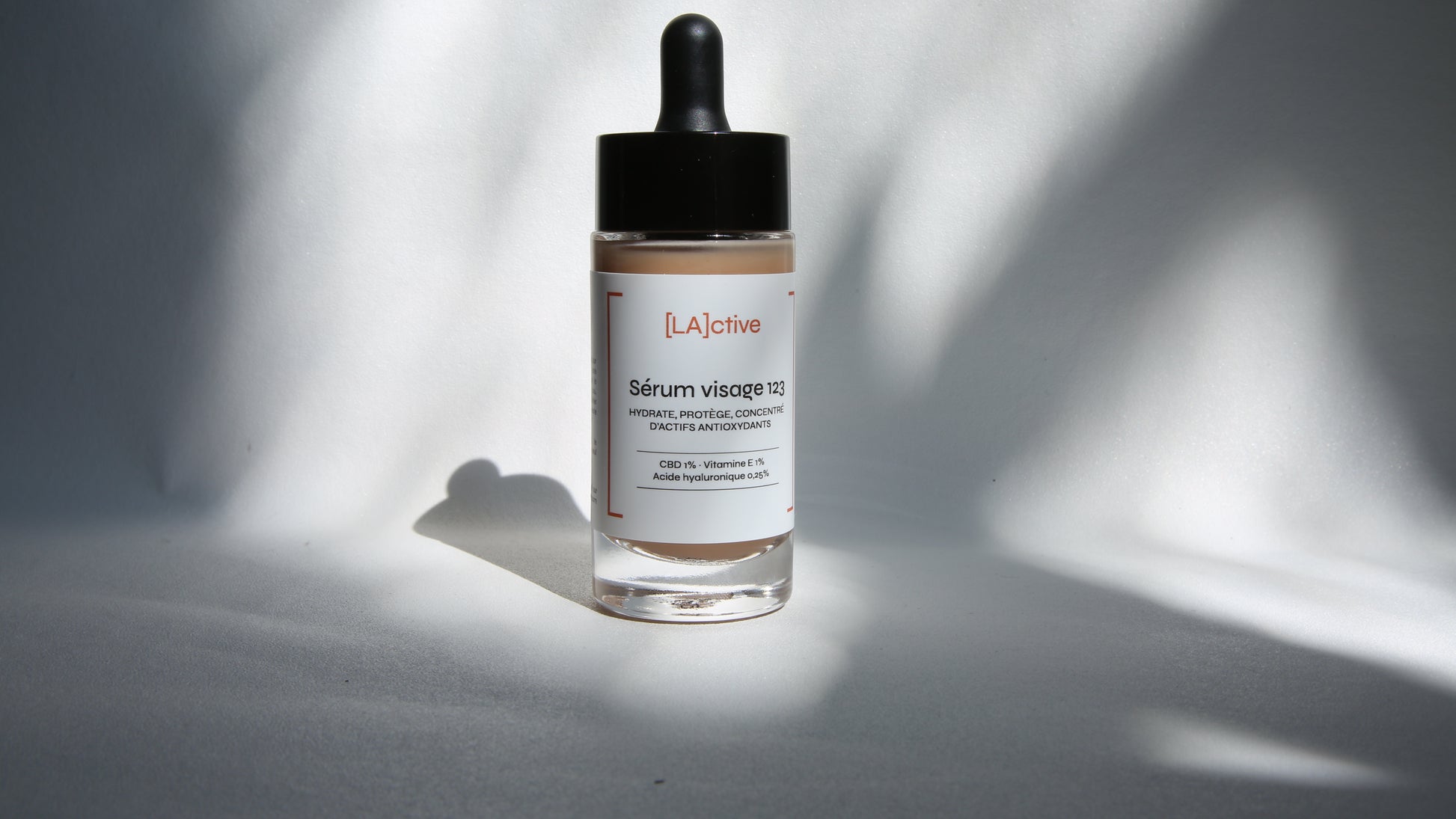 A bottle of facial serum labeled "[LA]active Sérum visage 123" in English and French, with active ingredients listed including CBD (cannabidiol), vitamin C and hyaluronic acid. The bottle features a black dropper cap and sits against a softly lit, neutral background.